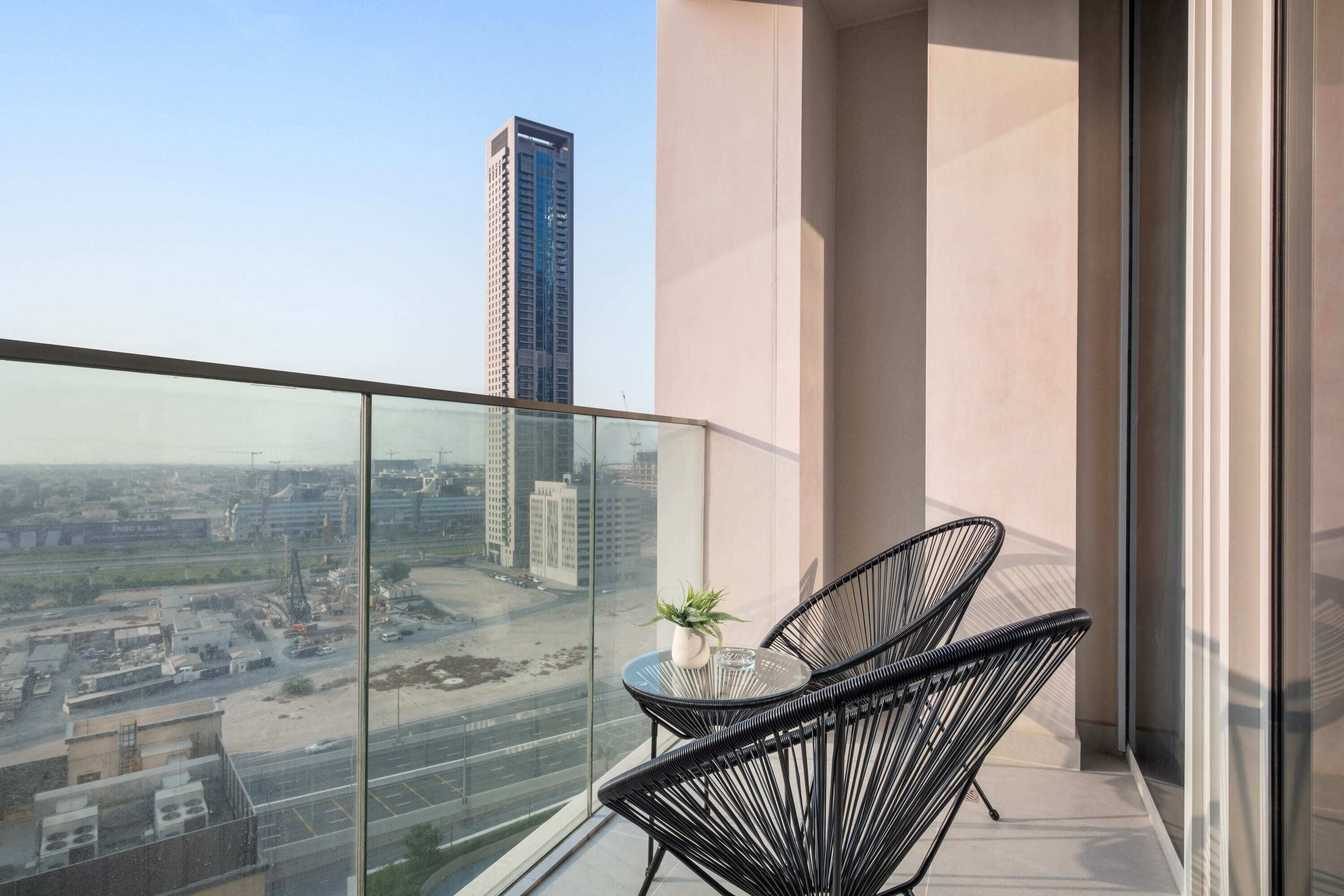 A balcony with a glass railing, two black wire chairs, a small table with a plant, and a view of a cityscape with buildings and a construction site.