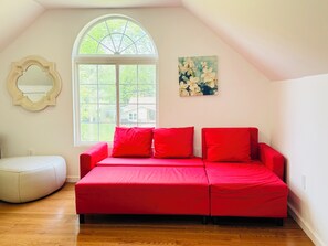 Um quarto acolhedor com um sofá vermelho, um banquinho branco, uma grande janela em arco e um quadro floral na parede.