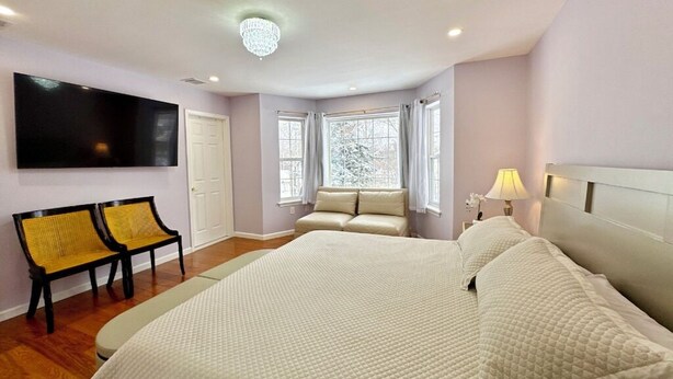A bedroom with a large bed, a TV mounted on the wall, a bench, and a window with curtains.