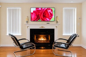 A living room with a fireplace, two leather chairs, a television on the wall, and white window blinds.