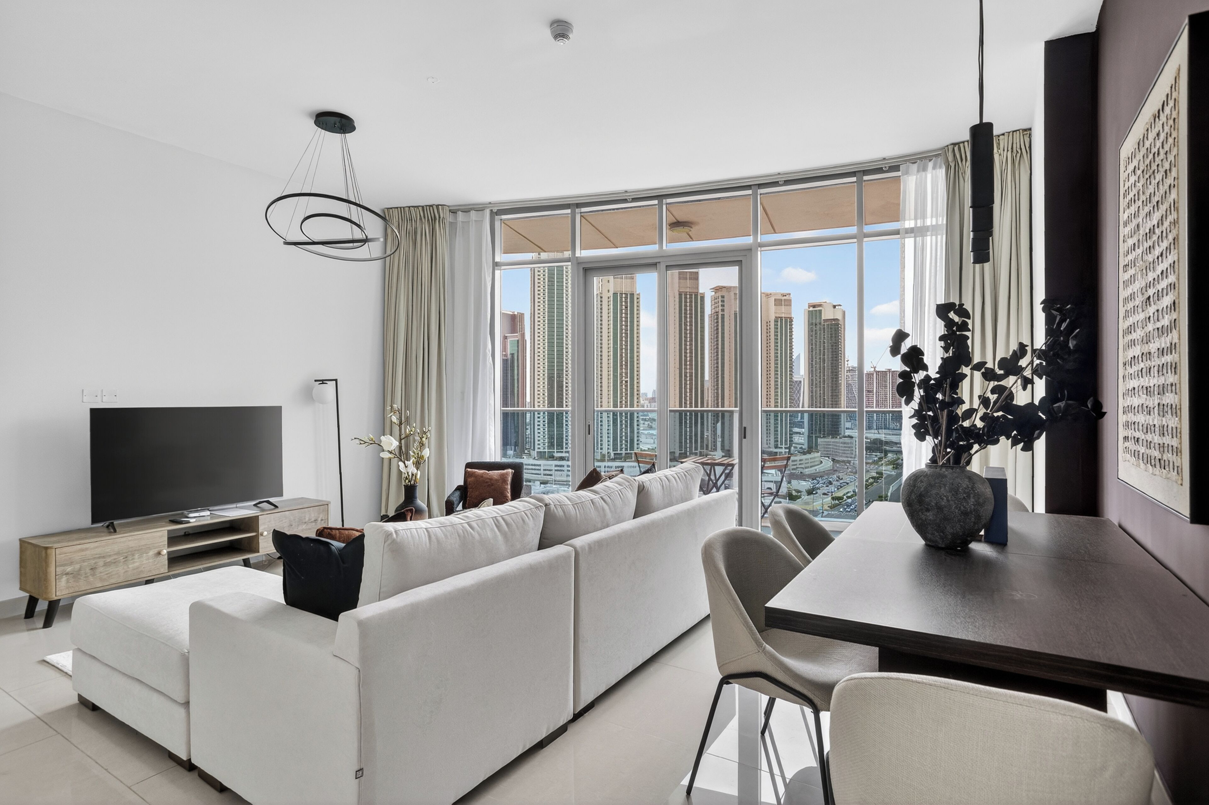 A modern living room with a large window, a flat-screen TV, a sofa, a dining table, and a vase with plants.