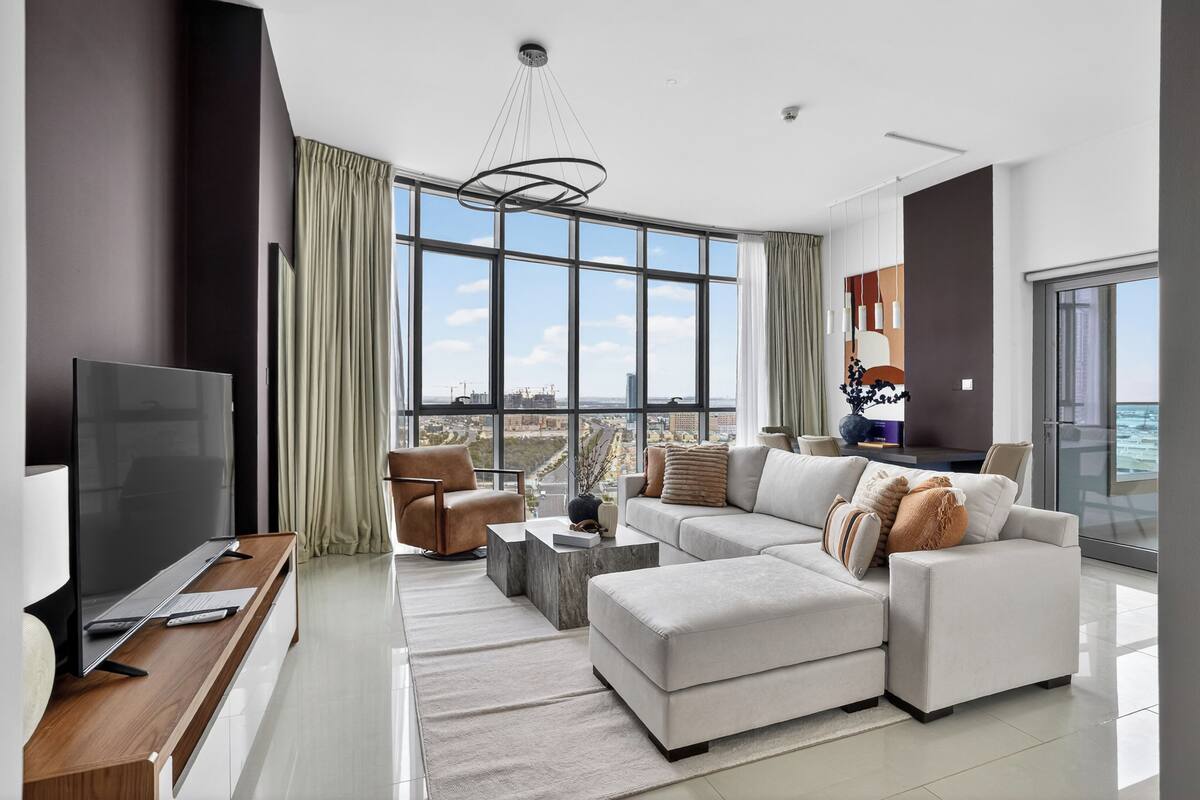 Un salon moderne doté d’un grand téléviseur, de sièges confortables, d’une table basse et offrant une vue sur le paysage urbain grâce à de grandes fenêtres.