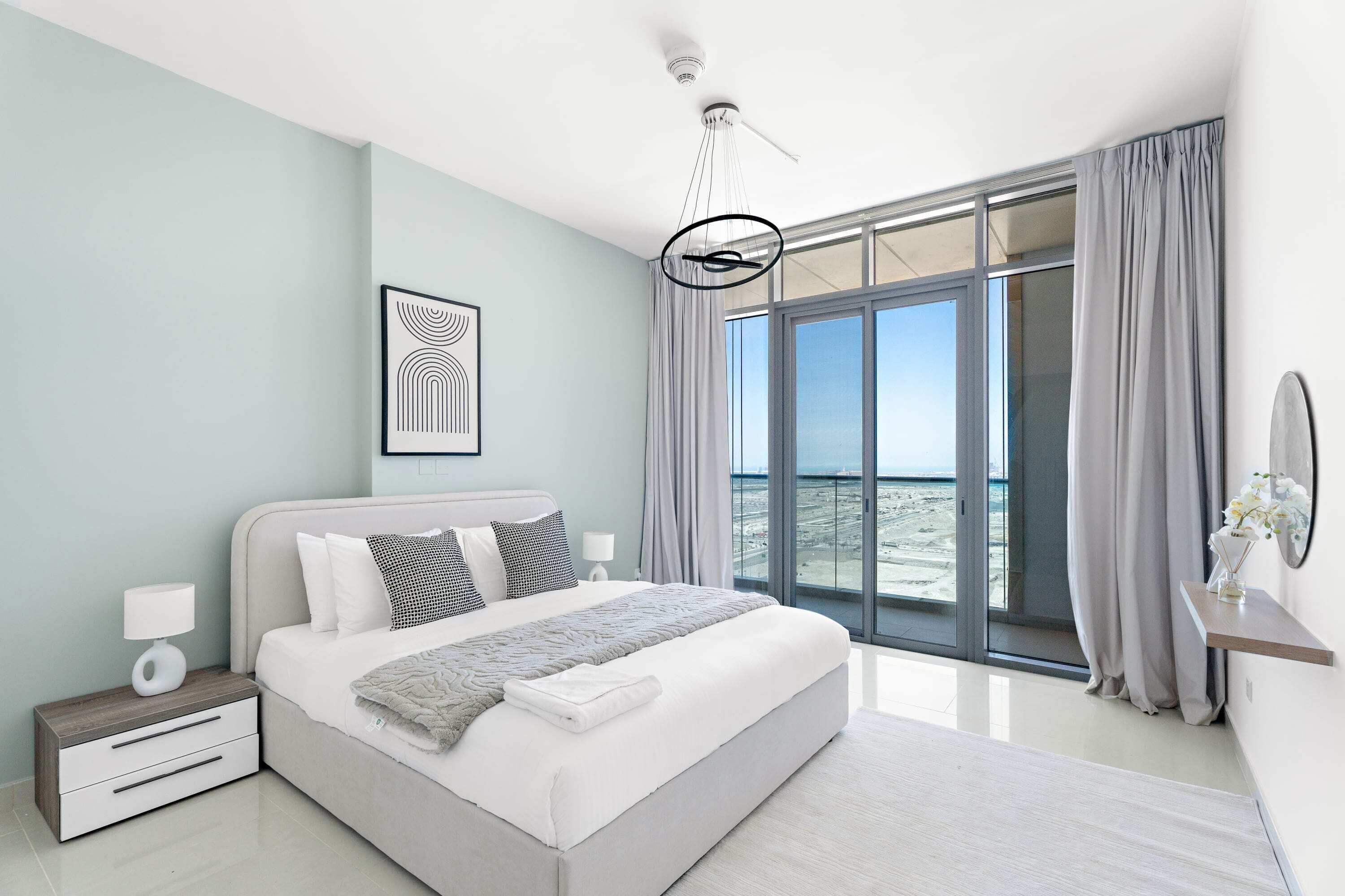 Een moderne slaapkamer met een groot bed, een nachtkastje, een bijzettafeltje met een lamp, een abstract kunstwerk in een lijst en uitzicht op het strand door grote ramen.
