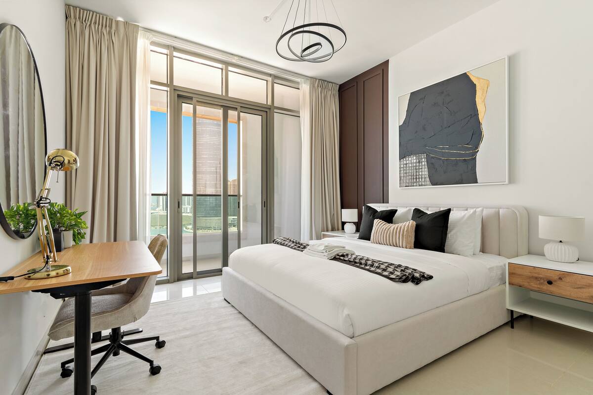 Une chambre d’hôtel moderne dotée d’un grand lit, d’un bureau, d’une chaise, d’une lampe, d’une table de chevet et d’une grande fenêtre avec des rideaux.