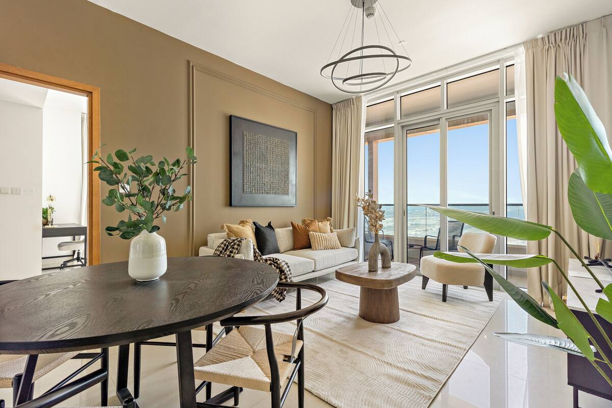 Un salon moderne avec une table à manger ronde, un canapé agrémenté de nombreux coussins, une table basse et une grande fenêtre donnant sur la mer.