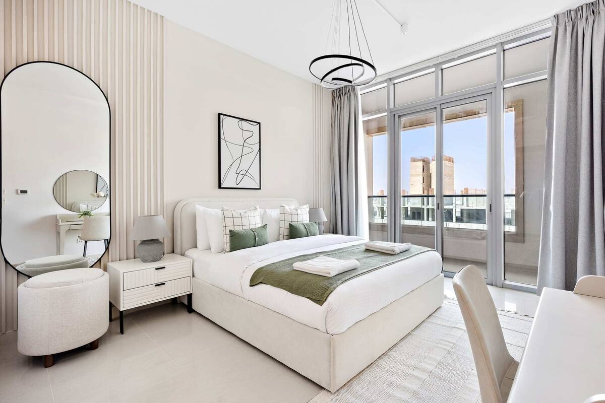 Une chambre d’hôtel moderne avec un grand lit, une table de chevet avec une lampe, un miroir et une vue sur la ville grâce à de grandes fenêtres.