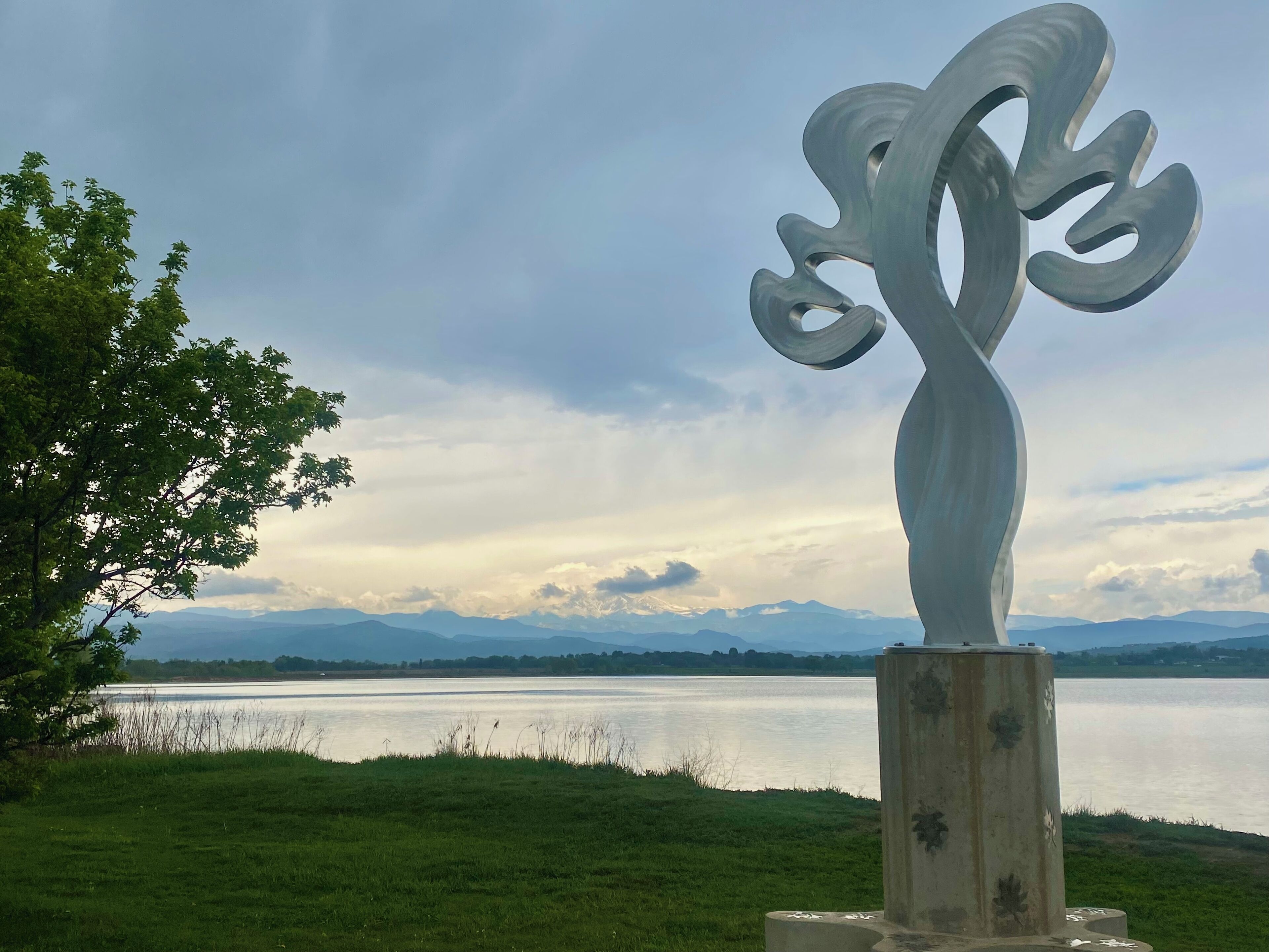 A sculpture of a stylized tree with a concrete base, set against a backdrop of a lake and distant mountains under a cloudy sky.