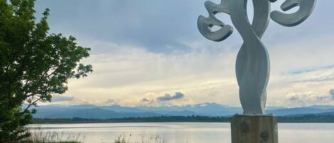 A sculpture of a stylized tree with a concrete base, set against a backdrop of a lake and distant mountains under a cloudy sky.