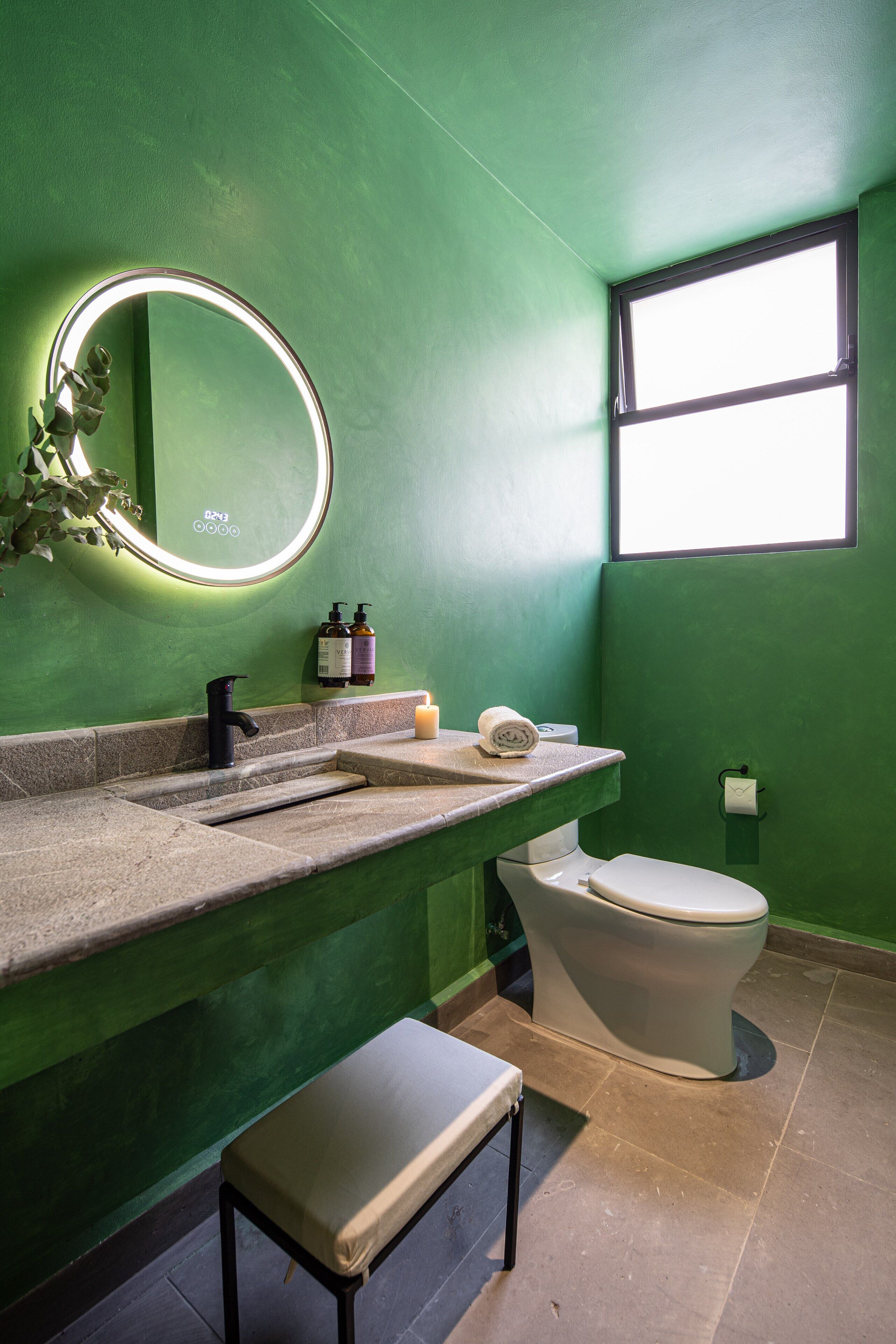 A bathroom with a green wall, a round mirror with lights, a sink, a toilet, and a bench.