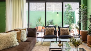 Una sala de estar acogedora con un ventanal grande, un sofá marrón, dos sillones, una mesa de centro de vidrio y varias plantas en macetas.