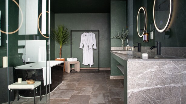 Un baño moderno con un lavamanos de mármol, dos espejos grandes con marcos iluminados, un perchero con batas blancas, un taburete pequeño y una planta en maceta.