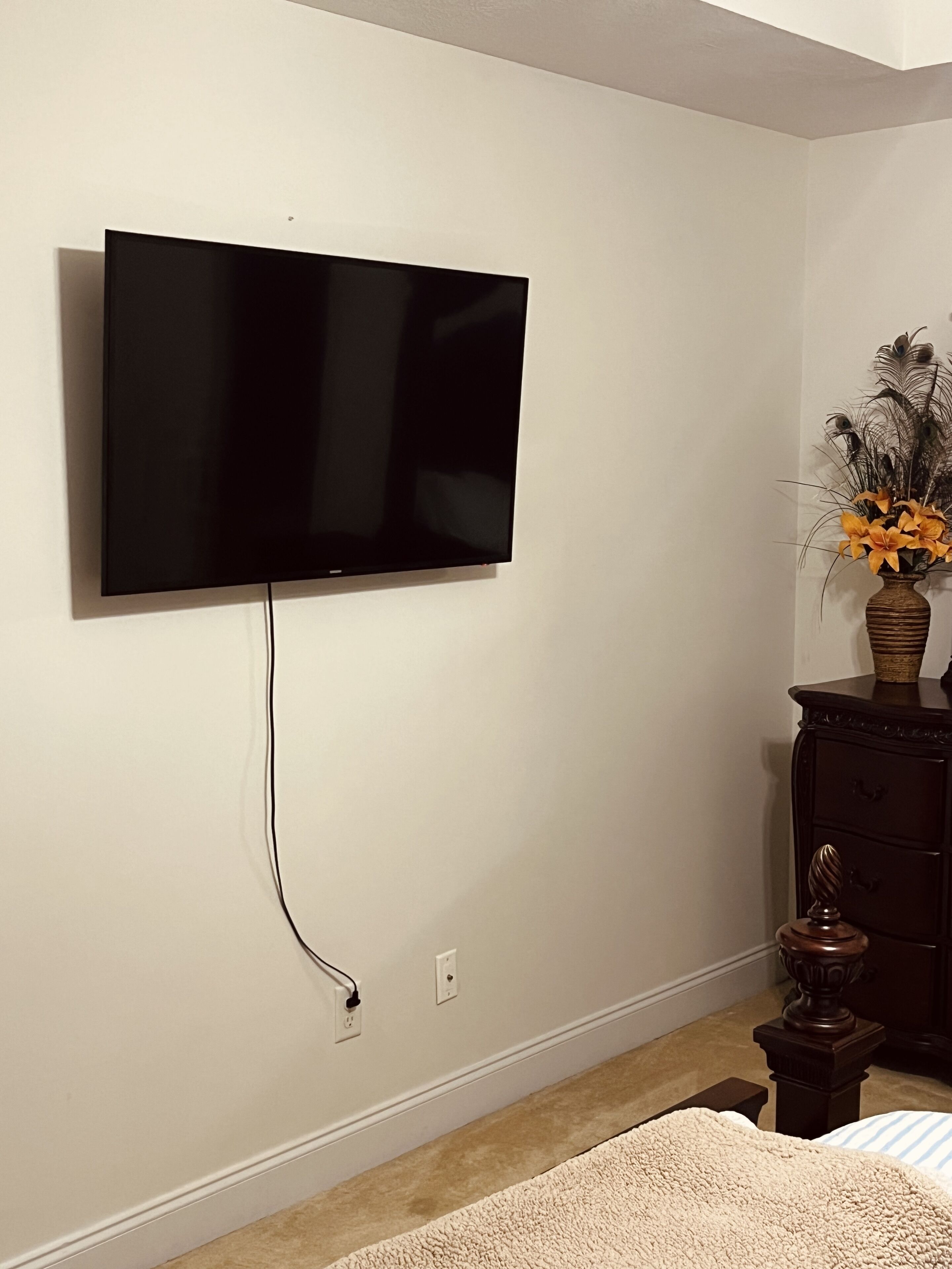 A room with a mounted flat-screen TV on the wall, a dark wooden dresser with a vase of flowers, and a beige carpet.