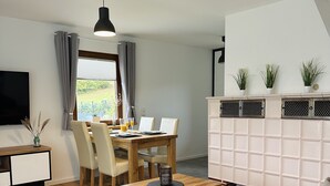 Ett modernt vardagsrum med en soffa, ett soffbord, ett matbord och stolar. Det finns en tv, ett skåp med växter och ett fönster med gardiner.