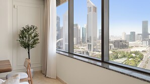 Une chambre moderne dotée d’une grande fenêtre donnant sur un paysage urbain, d’une plante en pot et d’un coin salon avec une chaise en bois et un siège rembourré.