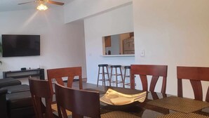 A living room with a sofa, a TV, a dining table with chairs, a plant, and a kitchen area in the background.