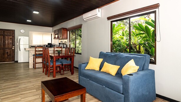 A living room with a blue sofa, wooden coffee table, and yellow cushions. There is a dining area with wooden chairs and a table. - Charming, beautiful cabin. (La Fortuna)