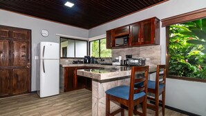 A kitchen with wooden cabinets, a refrigerator, a microwave, and a coffee maker. There are two wooden chairs with blue cushions. - Charming, beautiful cabin. (La Fortuna)