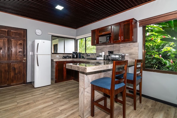 A kitchen with wooden cabinets, a refrigerator, a microwave, and a coffee maker. There are two wooden chairs with blue cushions.