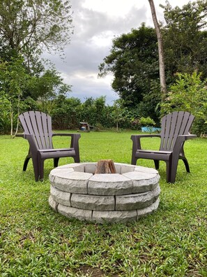 Two Adirondack chairs facing a stone fire pit in a grassy outdoor area with trees in the background.