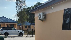 Una piscina con un borde de mosaicos hexagonales, un coche blanco aparcado cerca y un edificio con fachada color crema y un equipo de aire acondicionado instalado en la pared.