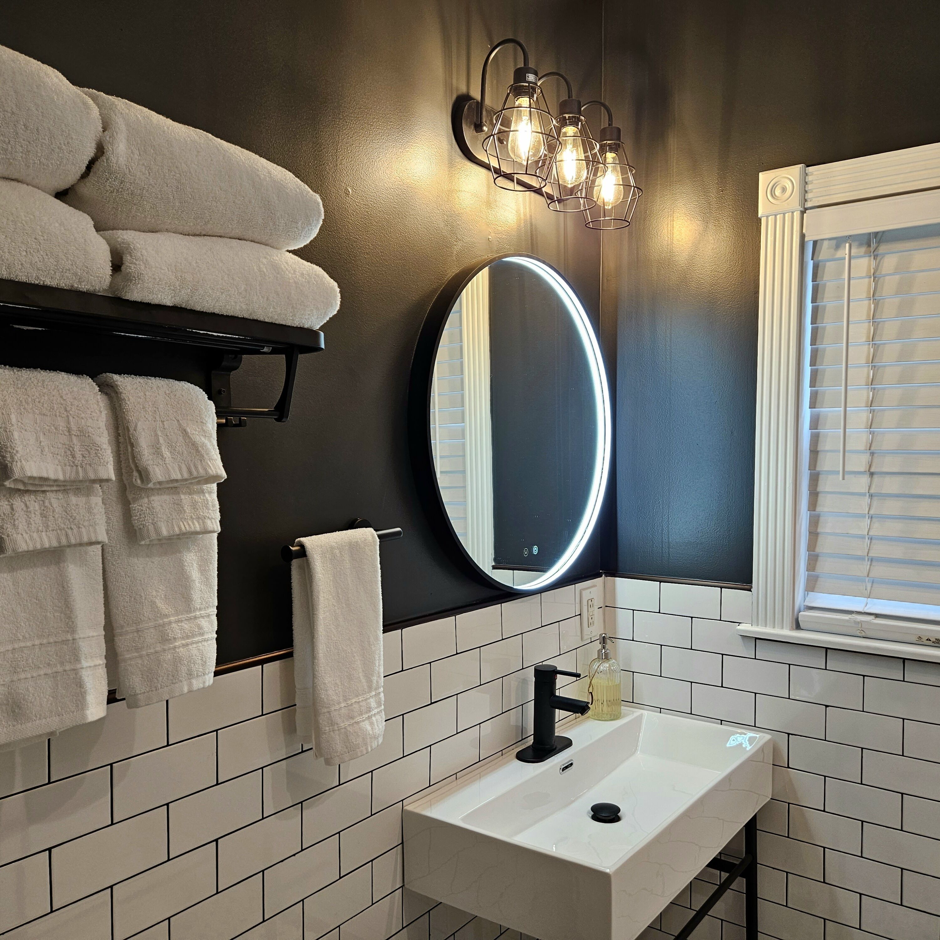Un baño con un lavabo blanco, un espejo con iluminación, un perchero con toallas y una ventana con persianas.