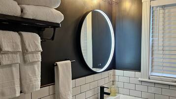 Un baño con un lavabo blanco, un espejo con iluminación, un perchero con toallas y una ventana con persianas.