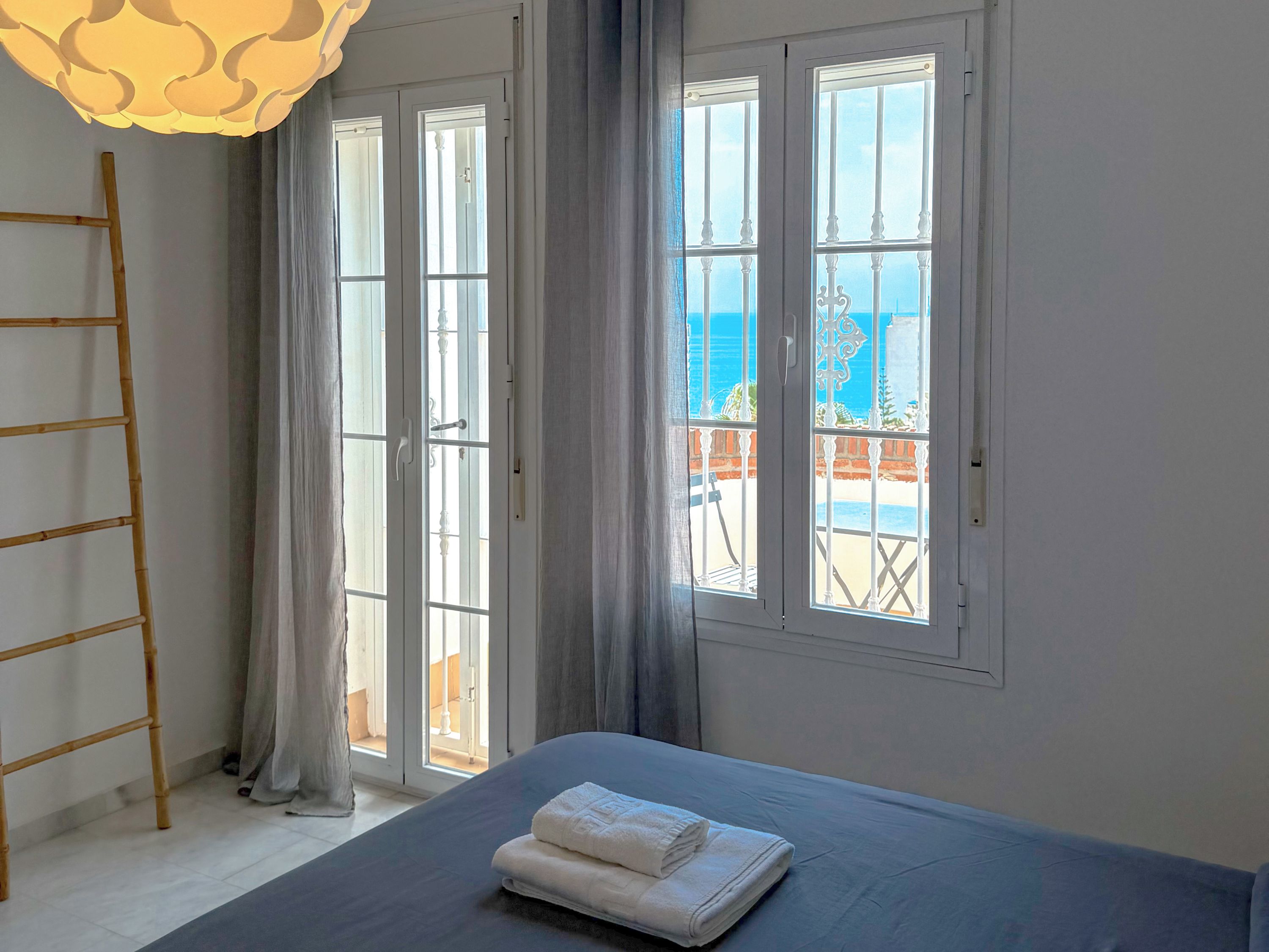 A bedroom with a bed, a wooden ladder, and a view of the sea through large windows.