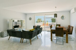 Un salon moderne comprenant un canapé, une table à manger et des chaises, un téléviseur à écran plat, et une vue sur la plage grâce à de grandes portes vitrées.