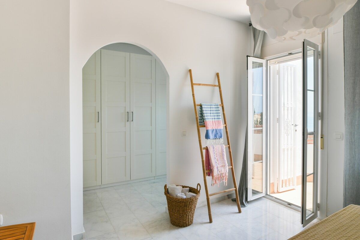 A room with a white wardrobe, a wooden ladder, a wicker basket, and a sliding glass door leading to an outdoor area.