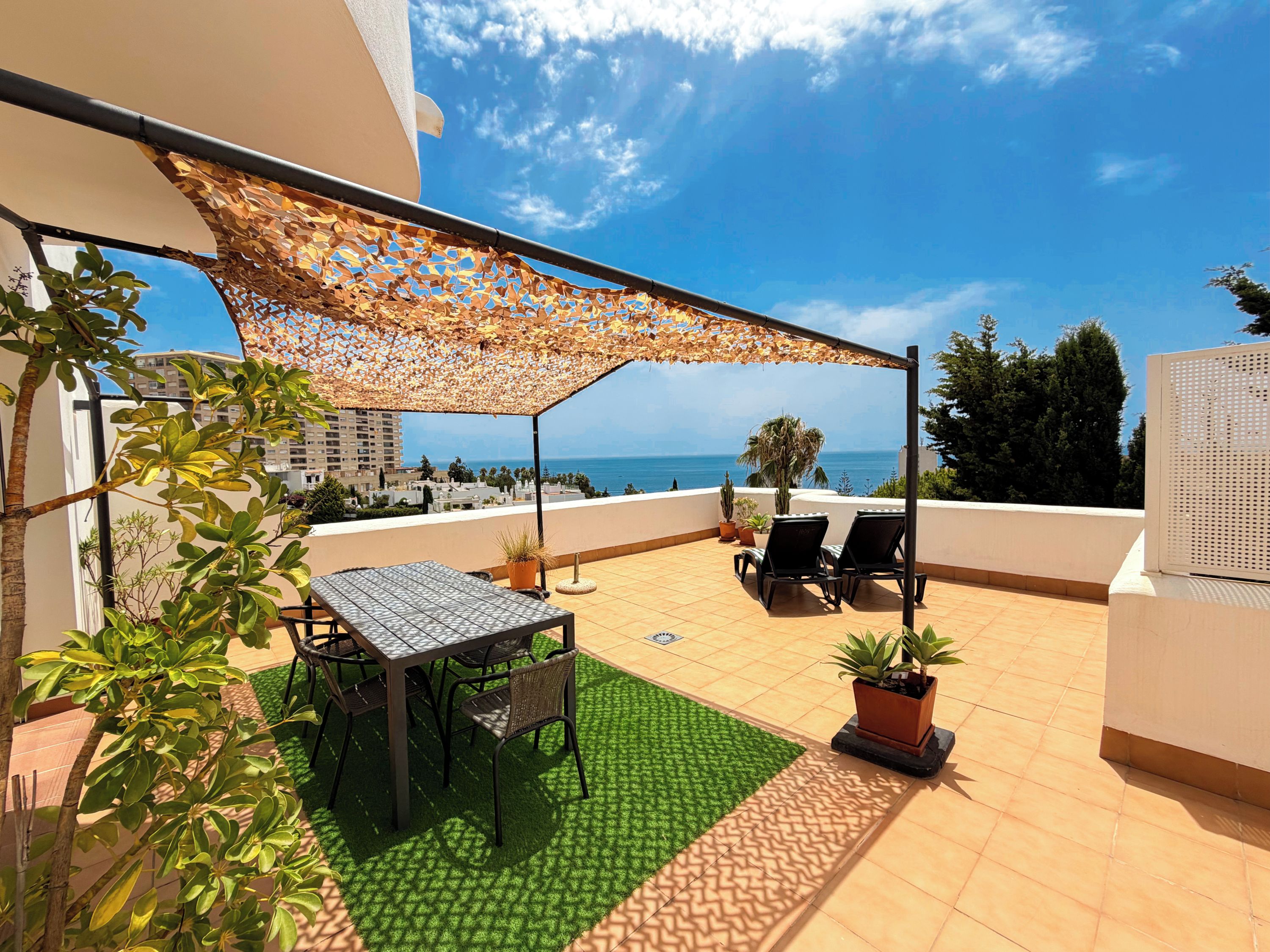 Una terraza en la azotea con mesa y sillas, una zona de césped artificial verde, plantas en macetas y vista al mar.