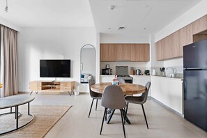 A modern living room with a flat-screen TV, a wooden dining table, and a sofa. The kitchen area includes wooden cabinets, a black refrigerator, and a microwave.