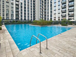 A swimming pool with metal railings, surrounded by a tiled deck and a high-rise building with balconies in the background.