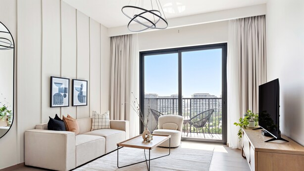 A modern living room with a sofa, coffee table, and TV, large windows with a city view, and a balcony with chairs. - Silkhaus Executive Residences, Dubai Hills (Dubai)