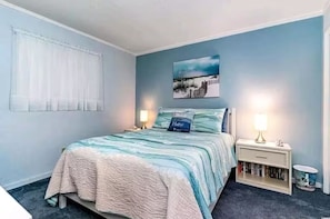A bedroom with a bed, two bedside lamps, a nightstand with a lamp, a wall-mounted picture, and a window with sheer curtains.