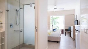 A hotel room with a glass-enclosed shower, a bed with a blue and white striped pillow, a desk with a chair, and a kitchenette.