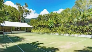 A tennis court with a net, surrounded by lush greenery and a building with a white roof.