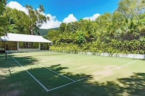 Tenniskenttä, jossa on verkko, ja jonka ympärillä on vehreää kasvillisuutta sekä valkoisen katon omaava rakennus.