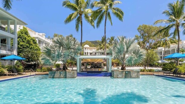 A swimming pool with lounge chairs and umbrellas, surrounded by palm trees and a building with balconies.