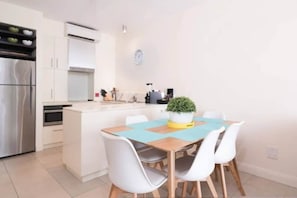 Une cuisine moderne avec des meubles blancs, un réfrigérateur en acier inoxydable et un coin repas comprenant une table en bois et des chaises blanches.