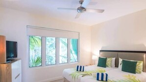 A hotel room with a bed, a TV, a ceiling fan, and a window with a view of greenery.