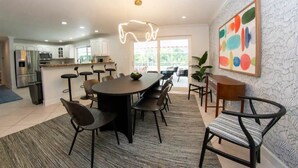Cozinha moderna com armários brancos, área de refeições com mesa e cadeiras redondas, e uma janela grande que ilumina com luz natural.