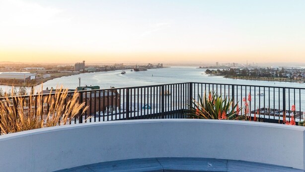 Un balcon donnant sur le fleuve, avec une balustrade, d’où l’on peut admirer des bateaux et, au loin, des bâtiments industriels.