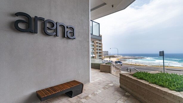 Un bâtiment moderne, avec une enseigne indiquant « arène », un banc, un bac à fleurs orné de plantes et une vue sur l’océan et la plage.