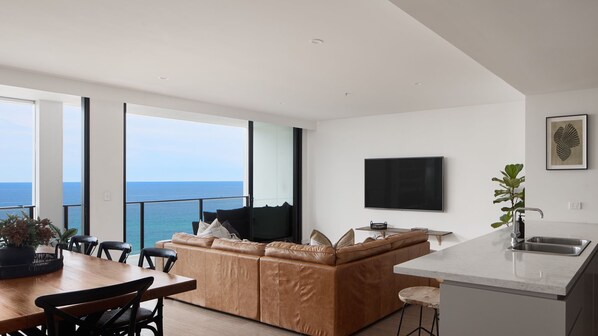 Un espace de vie moderne doté d’une grande fenêtre donnant sur la mer, d’un téléviseur à écran plat fixé au mur et d’un coin cuisine avec un évier et un plan de travail.