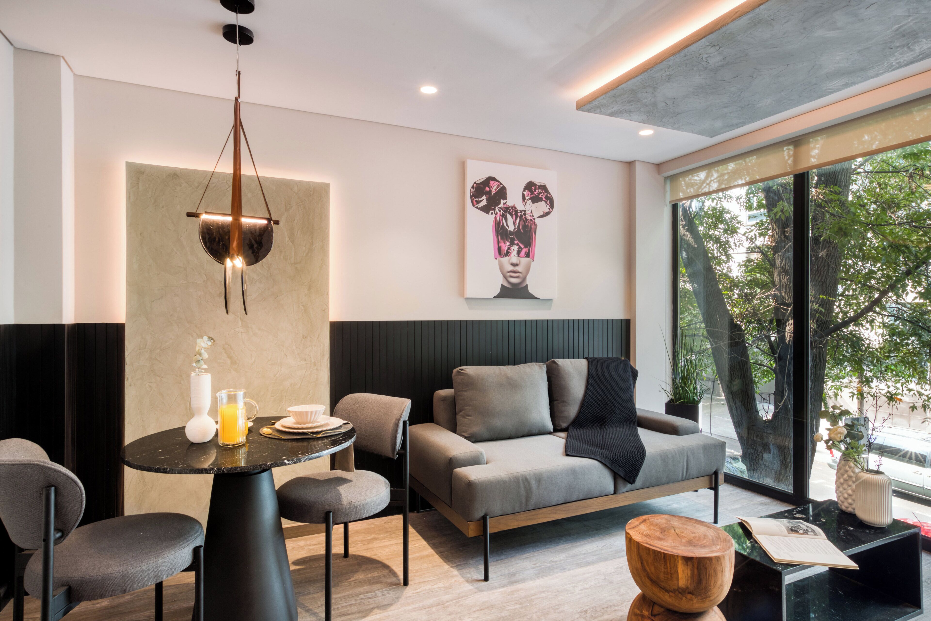 A modern living room with a sofa, coffee table, and dining area. There is a large window providing a view of trees and a street.