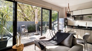 Una cocina moderna con encimera negra y electrodomésticos de acero inoxidable. Hay una zona de comedor con mesa y sillas, y un amplio ventanal corredero que da a un patio con plantas y una silla.