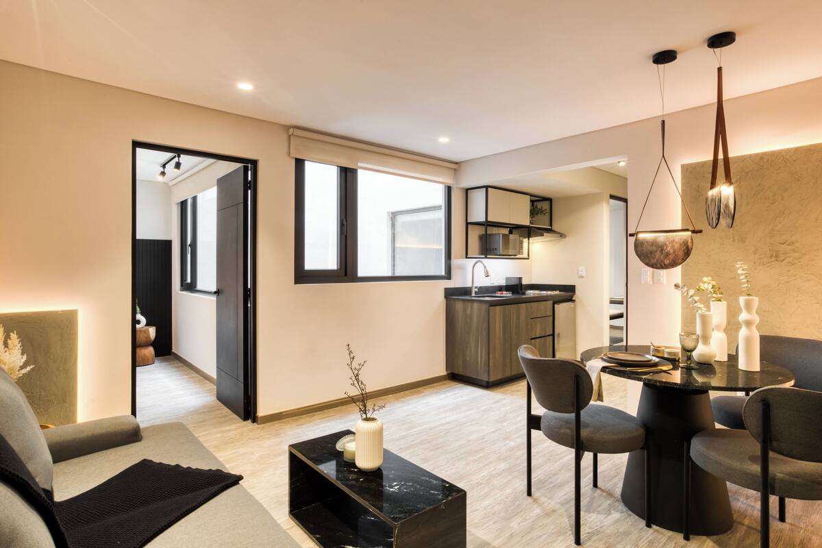 A modern living area with a sofa, a coffee table, and a dining table with chairs. There is a kitchen in the background with a microwave and cabinets.