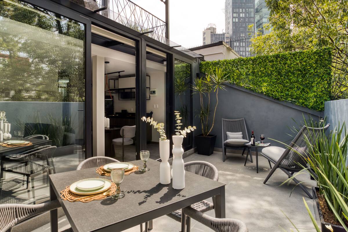 An outdoor patio with a dining table set for two, surrounded by wicker chairs. There are two white vases with orchids on the table.
