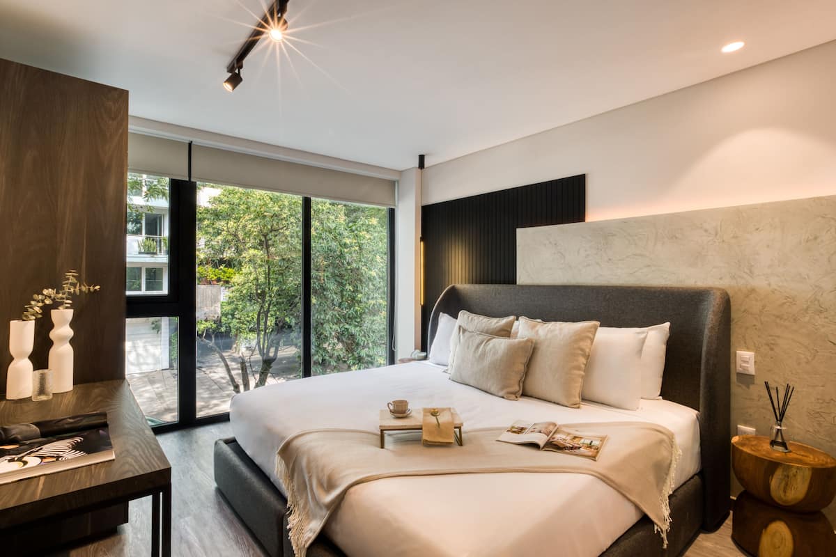 A modern bedroom with a large bed, a bedside table, a nightstand, and a view of greenery outside.