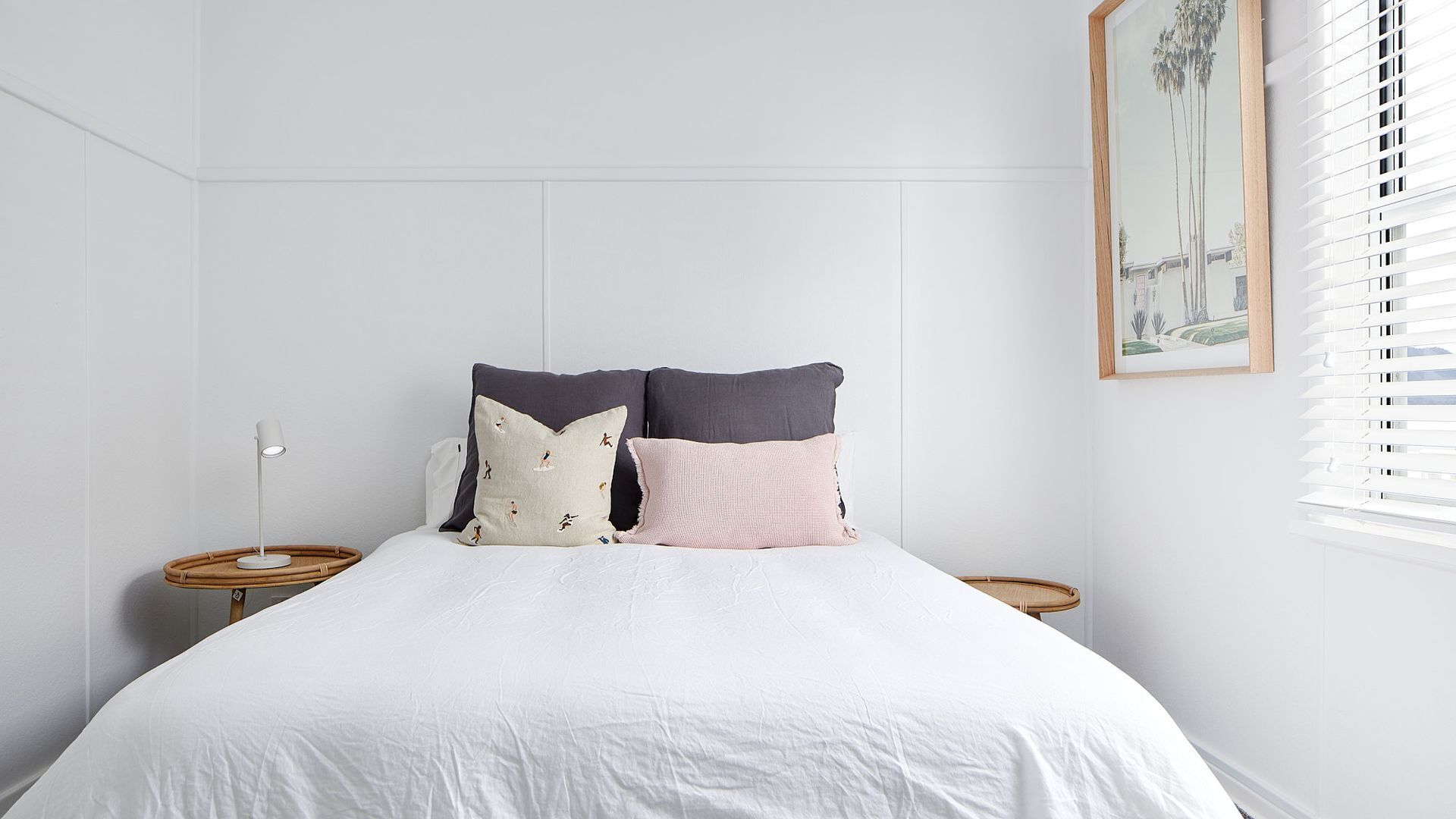 A bedroom with a white bed, two bedside tables, and a window with blinds.